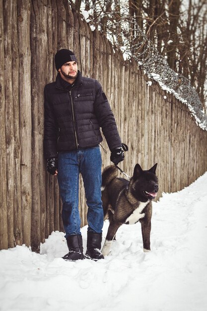 Een man in een jas en een gebreide muts loopt door een besneeuwd bos met een Amerikaanse Akita-hond