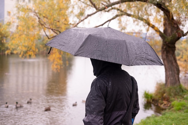 Een man in een jas met een capuchon staat met zijn rug met een paraplu in zijn handen Tegen de achtergrond van het meer