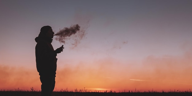 Een man in een jas rookt een elektronische sigaret buitenshuis Vapen op een zonsondergang op de achtergrond