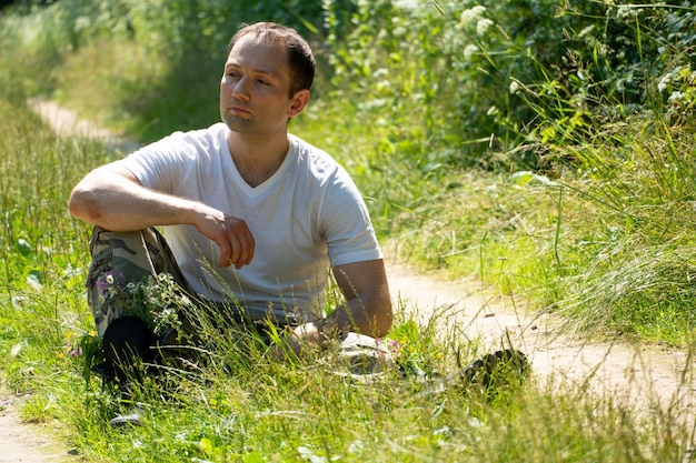 Een man in een militair uniform zit op de grond tegen de achtergrond van een onverharde weg Een jonge knappe man in een wit T-shirt ontspant in de natuur