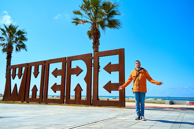 Een man in een oranje windjack poseert bij een enorme kunstinstallatie van de letters waar