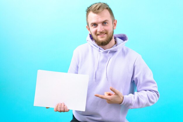 Een man in een paarse hoodie houdt een leeg wit A4-vel vast en wijst ernaar op een blauwe achtergrond Kaukasisch Tekst Knap Show Print Vriendelijk Document Mock Geluk Adverteren Portret Vrolijk