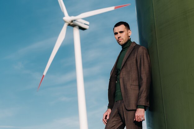 Een man in een pak met een groen golfshirt staat naast een windmolen tegen de achtergrond van het veld en de blauwe hemel. Zakenman in de buurt van de windmolens. Modern concept van de toekomst.