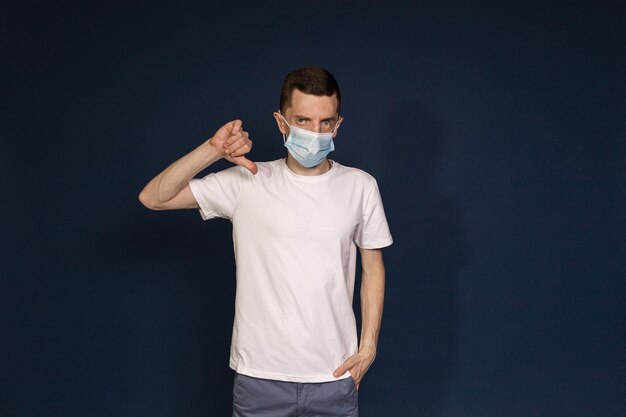 Een man in een wit T-shirt en een beschermend masker staat tegen een blauwe achtergrond en wijst met zijn duim naar beneden