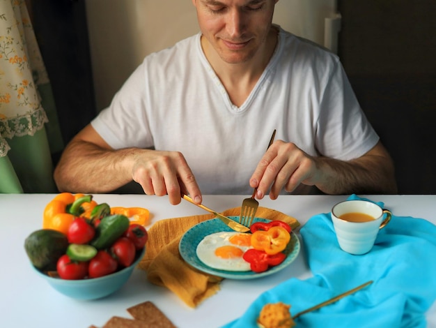 Een man in een wit T-shirt zit aan een tafel met een gezond ontbijt met eieren en groenten