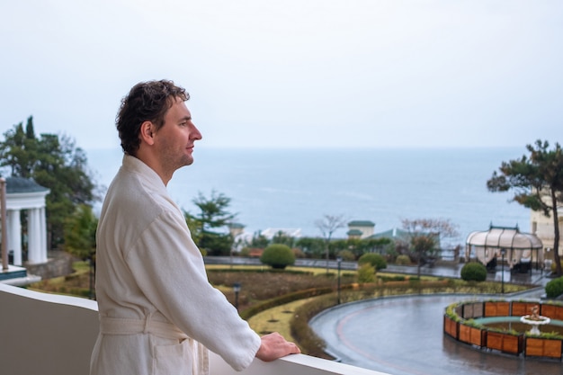 Een man in een witte badjas staat op het balkon van de residentie met uitzicht op de zee