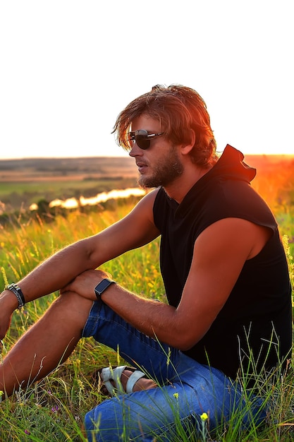 een man in een zwart t-shirt en een zonnebril zittend op het gras bij zonsondergang