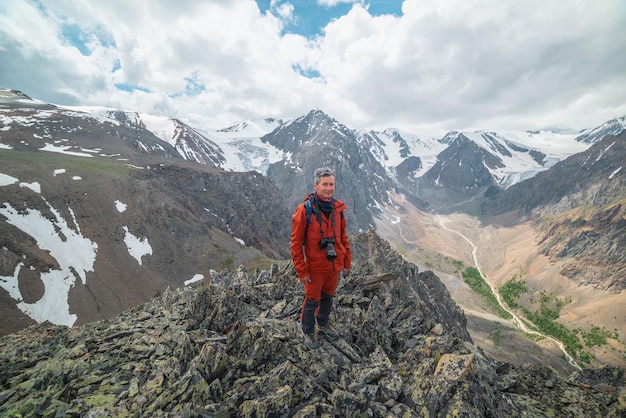 Een man in het rood met camera nabij de rand van de afgrond met scherpe rotsen met uitzicht op grote besneeuwde bergen en gletsjer onder bewolkte hemel Toerist op stenen heuvel met uitzicht op bergketen bij wisselvallig weer