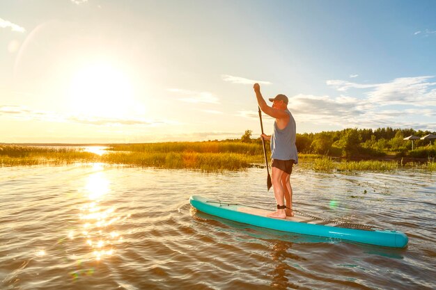 Foto een man in korte broek en een t-shirt op een sup-board met een roeispaan drijft op het water tegen de achtergrond van de ondergaande zon