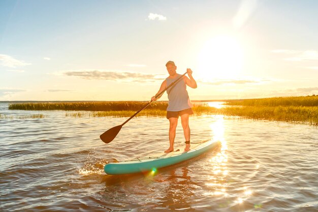 Een man in korte broek en een T-shirt op een SUP-bord met een roeispaan drijft op het water tegen de achtergrond van de zonsondergang