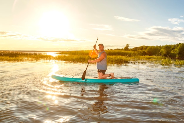 Een man in korte broek zit op een sup board bij zonsondergang in het water