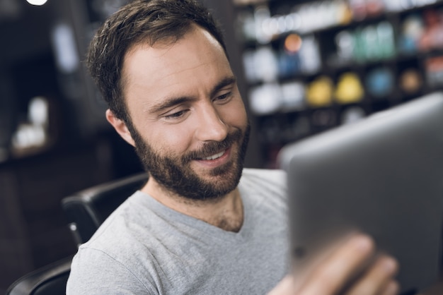 Een man kijkt naar tablet in de stoel van de kapper in de herenkapper.