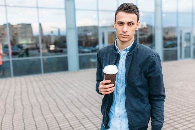 Een man loopt door de stad en drinkt koffie uit een papieren beker, een knappe man loopt rond en rust uit, de man op de achtergrond van een modern gebouw, kantoor