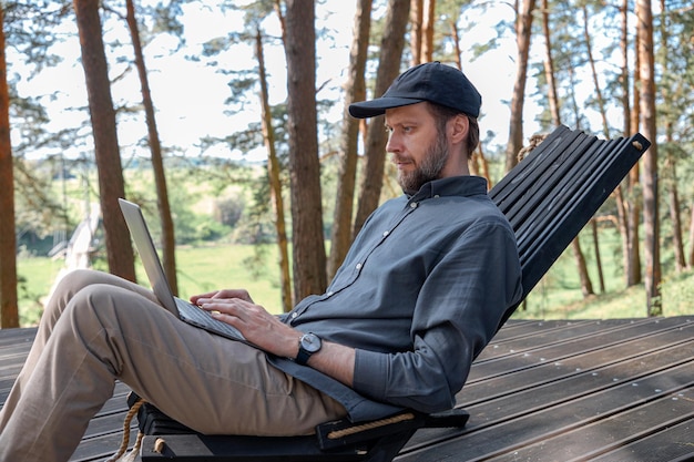 een man met een baard en een pet zit in de natuur en werkt op een laptop