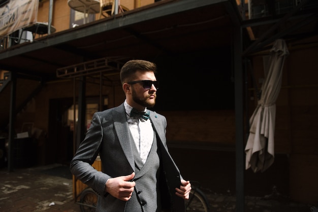 Een man met een baard en een zonnebril in een grijs pak poseert op straat om reclame te maken voor herenkleding.