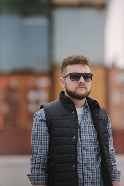 Een man met een baard en zonnebril in een geruite overhemd en vest poseren op straat