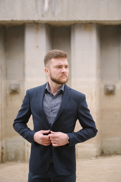 Een man met een baard in een blauw pak poseert op straat om reclame te maken voor herenkleding.