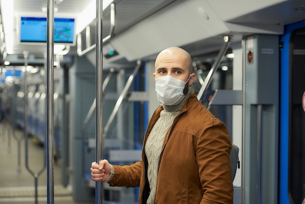 Een man met een baard in een medisch gezichtsmasker om de verspreiding van COVID-19 te voorkomen, staat en houdt de leuning vast in een metrowagen