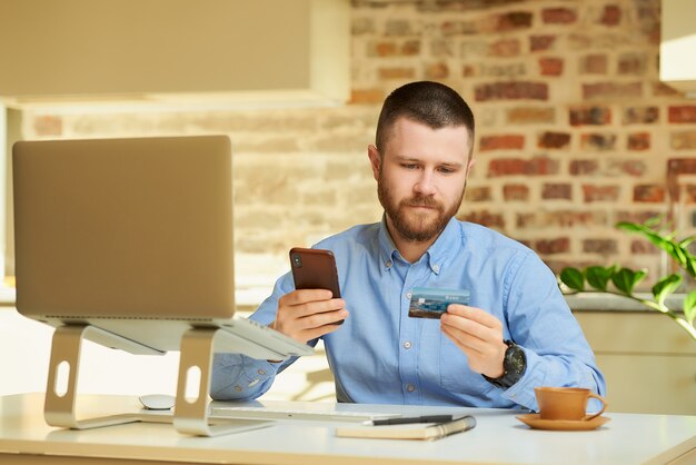 Een man met een baard leest informatie op de achterkant van de creditcard en typt deze op de smartphone om thuis een online aankoop te doen