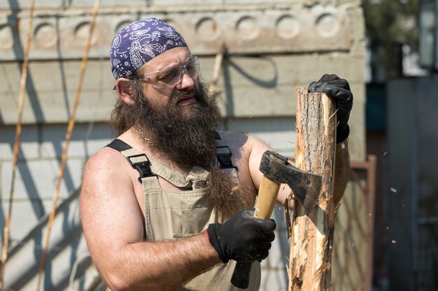 Een man met een baard van middelbare leeftijd in een bandana snijdt boomstammen met een bijl Brutaal in overall doet het harde werk