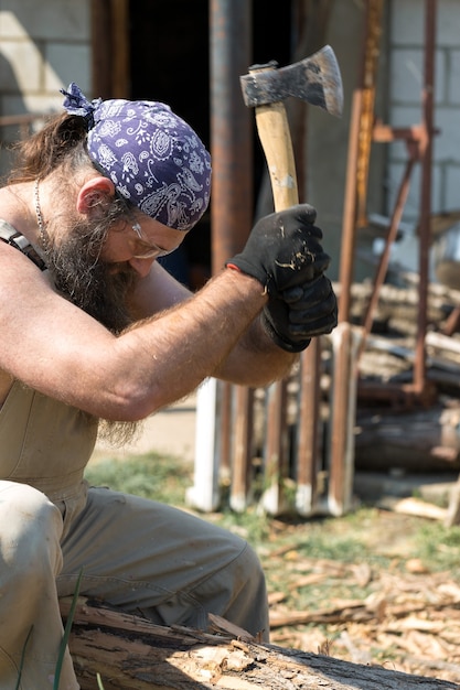 Foto een man met een baard van middelbare leeftijd in een bandana snijdt boomstammen met een bijl brutaal in overall doet het harde werk