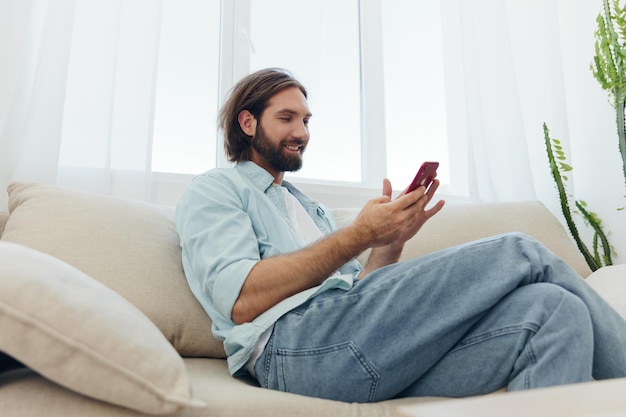 Een man met een baard zit overdag op de bank en glimlacht thuis en kijkt op zijn vrije dag naar zijn telefoon terwijl hij online op zijn telefoon sms't met vrienden