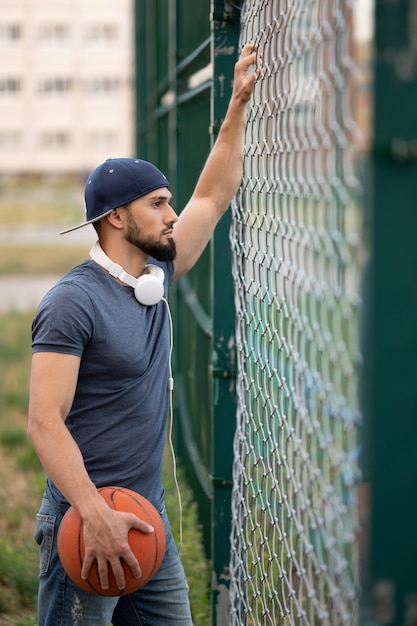 Een man met een basketbal kijkt overdag vanachter een hek op straat