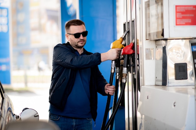 Een man met een blauwe jas en een donkere bril bij een tankstation. Hij vult de auto. Benzine. Levensstijl.