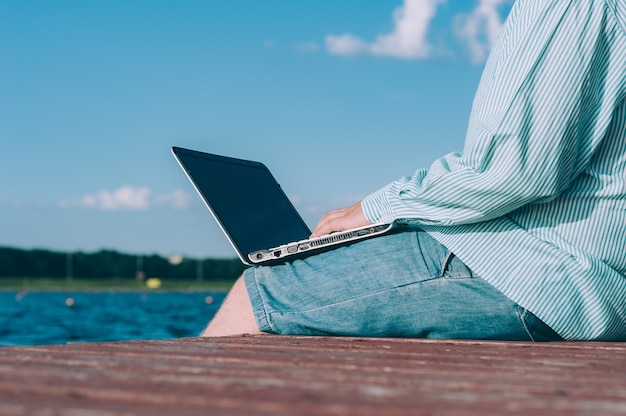 Een man met een bril en een overhemd werkt aan een laptop in de frisse lucht, tegen de ruimte van een rivier.