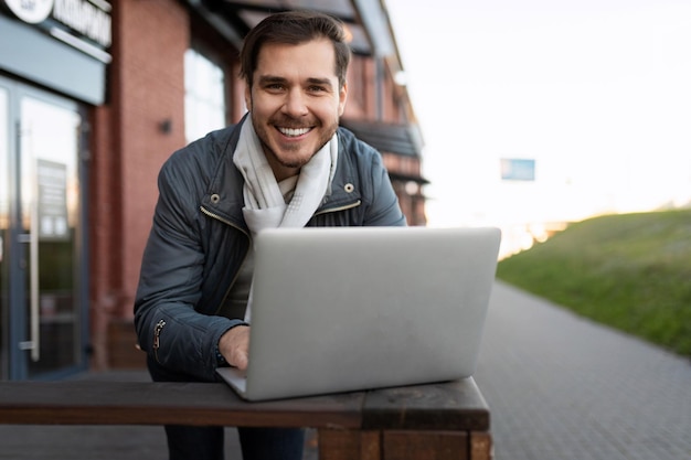 Een man met een glimlachende laptop kijkt breed naar de camera tegen de achtergrond van een stadsgebouw