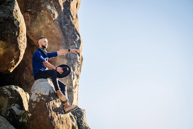 Een man met een hoed die hoog op de rotsen zit tegen de blauwe hemelachtergrond