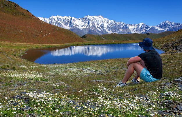 Een man met een hoed rust in een bloemenweide bij een meer met uitzicht op de met sneeuw bedekte bergen in Georgië Svaneti