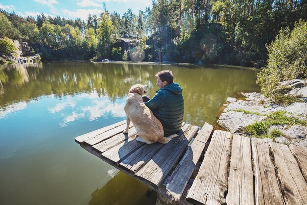 Foto een man met een hond zit op een houten terras aan een prachtige rotsachtige oever van een meer