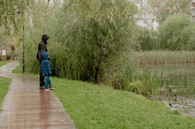 Een man met een kind in een jas met capuchon staat en kijkt naar het meer op een regenachtige dag