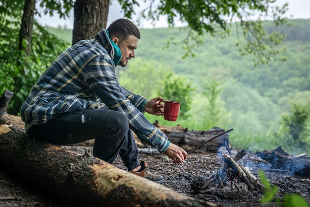 Een man met een kopje verwarmende drank warmt zich op bij het vuur in het bos.