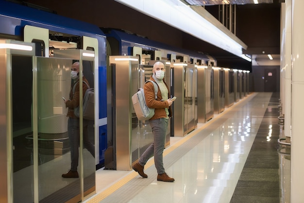 Een man met een medisch gezichtsmasker om de verspreiding van het coronavirus te voorkomen, houdt een mobiel vast terwijl hij een moderne metro verlaat