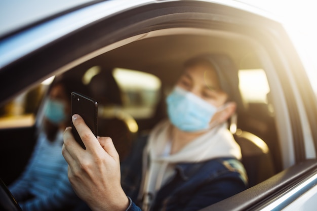 Een man met een medisch masker houdt een mobiele telefoon in zijn hand tijdens het autorijden