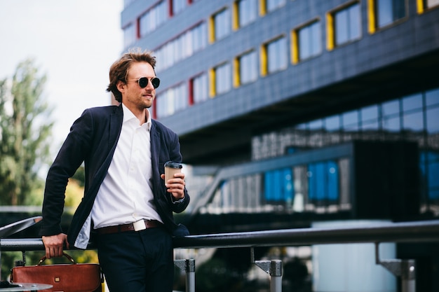 Een man met een mooie baard staat op straat in een blauw pak en een oranje lederen tas terwijl hij zijn koffie drinkt