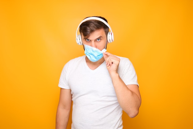 Een man met een muzikale koptelefoon en een medisch masker staat tegen een gele achtergrond.