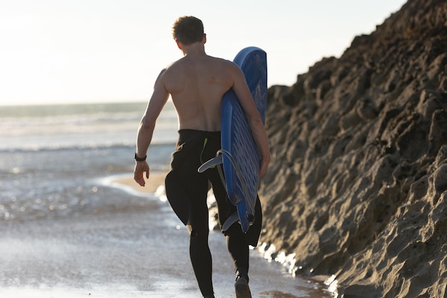 Een man met een naakte torso die bij een rots staat met een surfplank van achteren