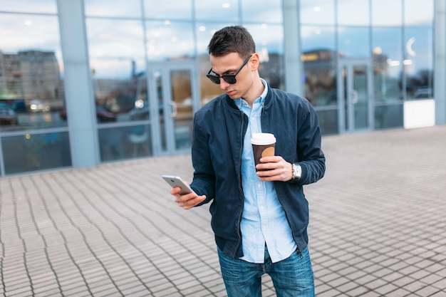 Een man met een papieren kopje koffie gaat door de stad, een knappe jongen in stijlvolle kleding en een zonnebril, die een telefoontje pleegt