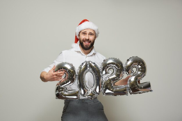 Een man met een roodfluwelen kerstmuts houdt zilveren ballonnen in de vorm van 2022 vast en doet wat. Een gelukkige man met een baard op een nieuwjaarsfeest.