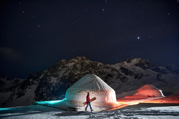 Een man met een snowboard die 's nachts in de winter in de bergen van Almaty Kazachstan in de buurt van een yurt loopt