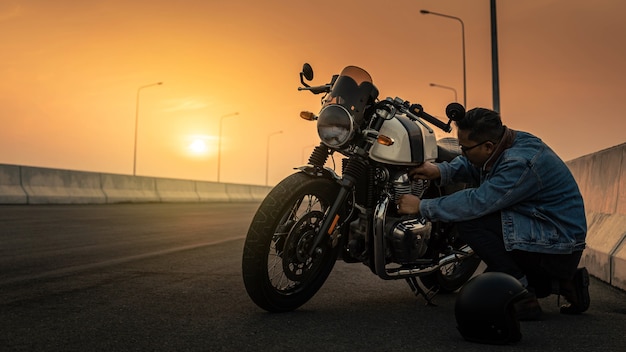 Een man met een spijkerjasje repareert een vintage motorbougie op een snelweg bij zonsondergang. Man rijden op een vintage motorfiets Biker Lifestyle Concept.