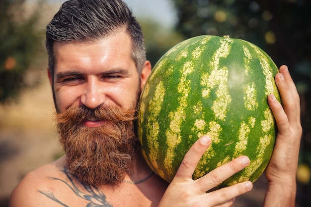Een man met een stijlvol kapsel, een lange baard en een tatoeage houdt een grote watermeloen en glimlacht
