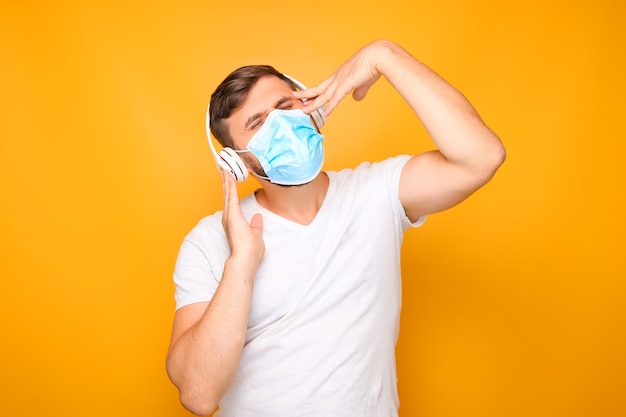 Een man met een witte muzikale koptelefoon staat op een gele achtergrond en draagt een medisch masker.