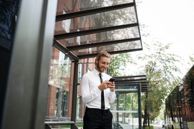 Een man met krullend haar, in een wit overhemd en een stropdas, bij de bushalte kijkt naar de telefoon terwijl hij op de bus wacht. het concept van een mobiele applicatie voor verkeer in de stad.