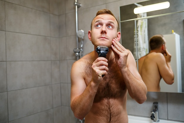 Een man scheert zijn gezicht met een elektrisch scheerapparaat voor een spiegel. Huidirritatie. Bad procedure