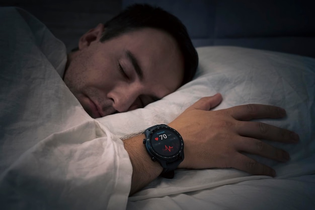 Een man slaapt op een bed met een hartslagcontrole in zijn slaap Slimme horloges meten de polsslag aan de hand van een slapende persoon Fitness Activity Tracker met hartslagfrequentie op de hand van de man boven het bed