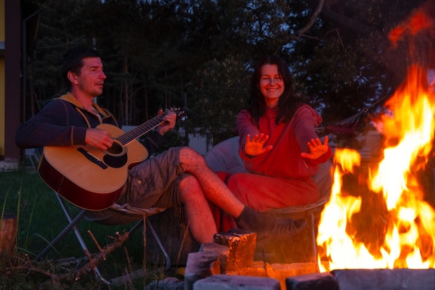 Een man speelt gitaar een vrouw luistert en zingt mee een verliefd koppel zit bij het kampvuur in de binnenplaats van het huis op campingstoelen een romantische avond